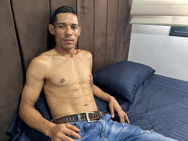 HarryTtaylor est un homme latino, musclé et torse nu, avec un corps mince et tonique et de petits tétons dressés, assis sur un lit avec des draps bleus, portant un jean déchiré et une ceinture marron..