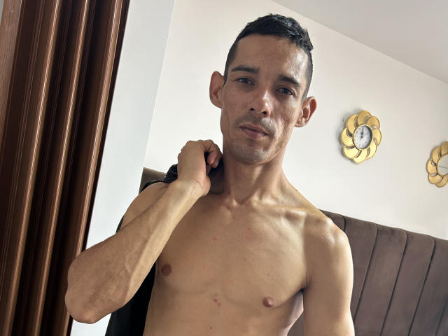 Harry Taylor, torse nu, tenant une sangle noire, a une silhouette mince et musclée, des cheveux courts et foncés, et un léger sourire. Ses petits mamelons flasques et sa poitrine nue sont visibles..