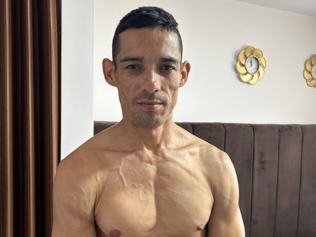 Harry Taylor sans chemise, avec une poitrine musclée, bronzée et légèrement poilue, des cheveux courts et foncés, et un sourire discret, debout devant un mur recouvert d'un tissu marron..
