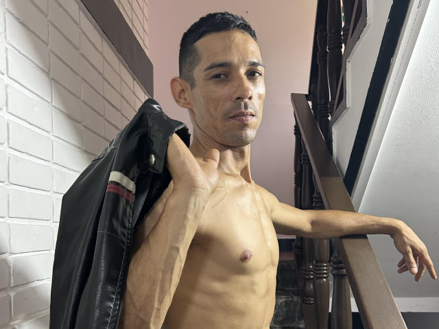 Harry Taylor sans chemise, avec une silhouette mince et tonique et de petites seins, tenant une veste noire sur son épaule, debout dans un escalier, regardant l'appareil photo..