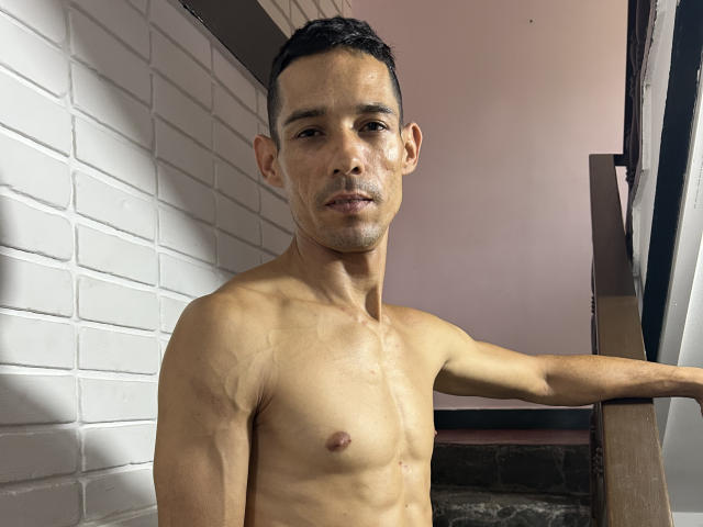 Harry Taylor, torse nu, avec une silhouette mince et musclée, de petites seins, des cheveux courts et foncés, et un léger bronzage, se tient debout avec une expression détendue et confiante, appuyé sur une rambarde en bois..