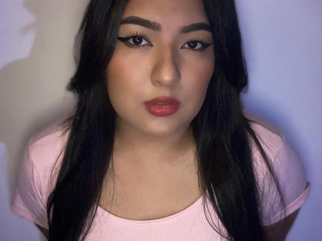 AuroraSxx with long black hair, full red lips, and winged eyeliner, wearing a pink shirt, looking directly at the camera with a neutral expression.