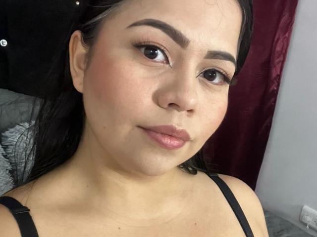 LilyRousee has light brown skin, dark eyebrows, and black eyeliner. She's wearing a black strap top with pinkish lips and a slight smile. Her ass is not visible.
