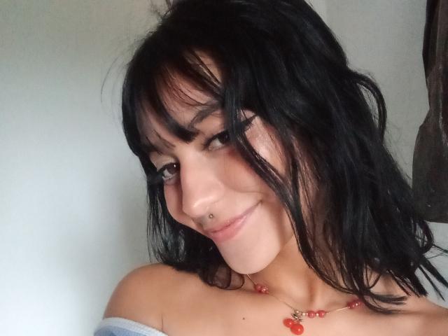 LindaWatsong with black wavy hair, smiling, has a nose piercing and wears an off-shoulder top, red bead necklace, and a subtle smirk. Her tits are partially visible.