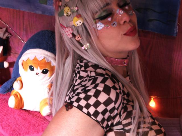 Toukauwu with silver hair, checkered shirt, pink collar, and glittery face paint, pouting with red lipstick, next to a chubby orange and white cat plushie.