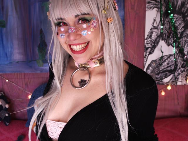 Toukauwu with white hair, colorful face stickers, black cardigan, pink lace bra, and a pink collar with a ring, smiling at the camera. Boobs visible, cute, sexy.