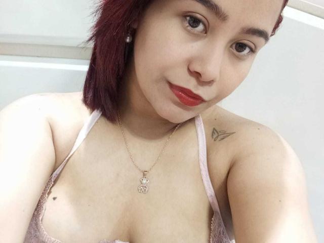 SalmaTorres, avec des cheveux roux, une peau claire, de petites seins, un haut en dentelle rose, du rouge à lèvres rouge, un collier, un tatouage sur l'épaule droite, souriant à la caméra..
