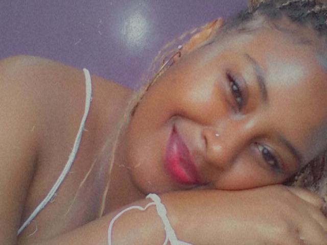 ChristyLeaX with a cute smile, pink lipstick, and braided hair, resting her head on her arm, wearing a white strap top, showing her tits. Soft, warm lighting.