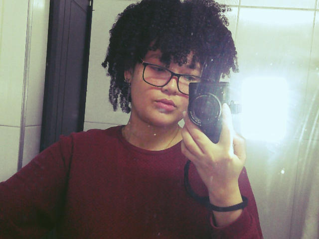 IrySunshine, avec ses cheveux noirs bouclés, ses lunettes et son pull rouge, tenant un appareil photo devant un miroir de salle de bain, le flash se reflétant, un bracelet noir à son poignet..