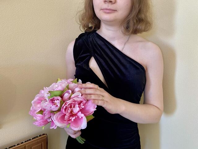 Steisii with light skin and wavy brown hair, wearing a black one-shoulder dress, holding a pink bouquet of peonies, standing against a beige wall.