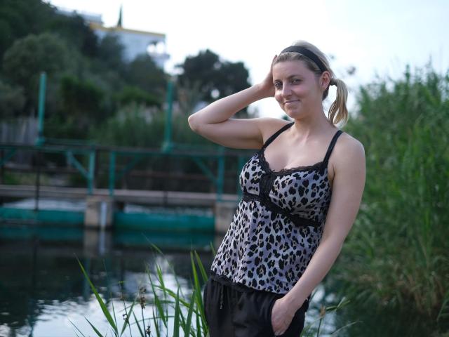 LoucilleDiamond-hot with small boobs, wearing a leopard-print tank top, black lace trim, and black pants, standing by a waterway, hand in hair, greenery in background, smiling slightly.