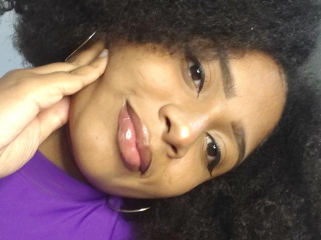 MeganWilliams has dark brown skin, curly black hair, and is sticking out her pink tongue. She's wearing a purple shirt, with a hand touching her cheek.