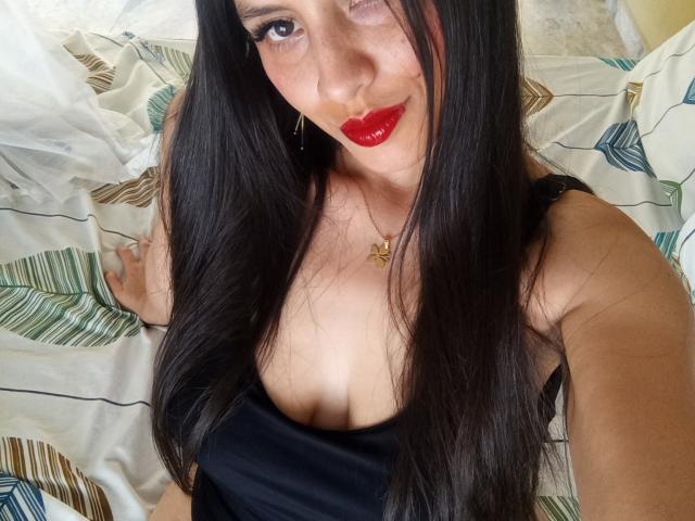 MadissonFontaine has long black hair, red lipstick, and a black tank top showing her tits, with a gold necklace and one eye winking. She's on a bed with patterned sheets.