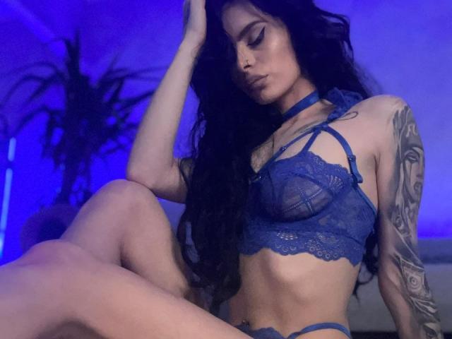 A DidiDancer, com longos cabelos pretos, vestindo lingerie azul de renda, está sentada com uma mão no cabelo, mostrando tatuagens no braço direito e um abdômen plano e tonificado. Suas nádegas estão visíveis..