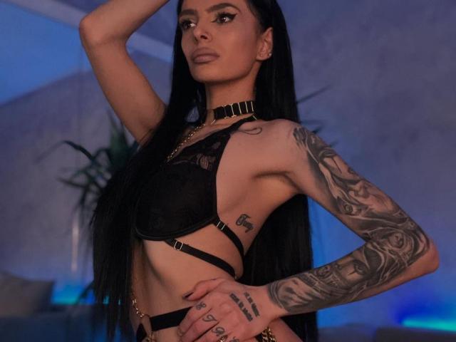 DidiDancer, com longos cabelos pretos, vestindo um sutiã de renda preto, um colar e roupa íntima de tiras, mostrando tatuagens no braço e no lado do corpo, com a mão na cabeça, com uma expressão confiante. Seus seios são pequenos..