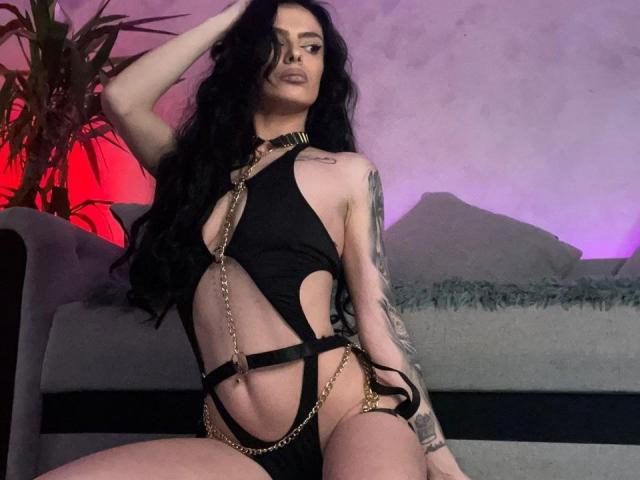 DidiDancer, com longos cabelos pretos, vestindo um conjunto de lingerie preta com tiras e correntes douradas, sentada em um sofá cinza, mostrando tatuagens em seu braço e olhando para a câmera..