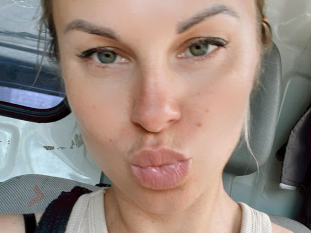BelleLisaG, con labios rosados y sensuales, ojos verdes y cabello castaño ligeramente despeinado, está en un coche, vestida con una camiseta sin mangas color beige, y hace una mueca de beso. Su nariz es pequeña y rosada..