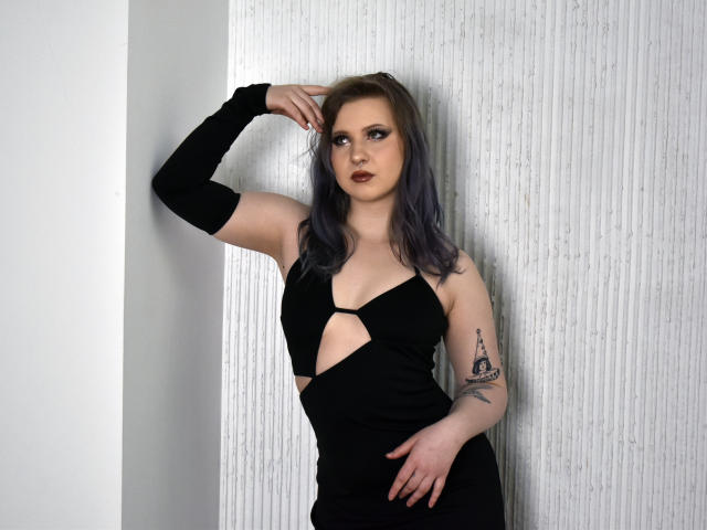 UrsulaAnderson with grey-blue hair, wearing a black cut-out dress, arm raised, exposing a tattoo on her left arm, standing against a white textured wall, looking at the camera.