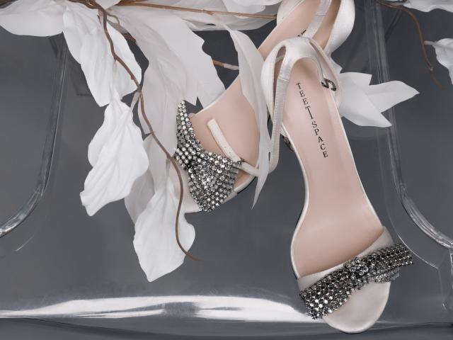 JeanieCat's white high-heeled shoes with shiny silver studs, criss-cross straps, and "Sofitale" label, resting on a reflective surface with white floral leaves.