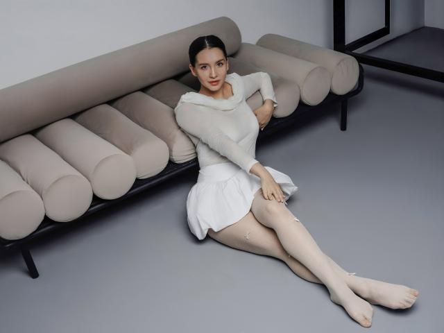 JeanieCat, with pale skin and dark hair, sits on a beige, cylindrical couch, wearing a white, long-sleeve, dress, and sheer tights, showing her bare, small-ass and feet.