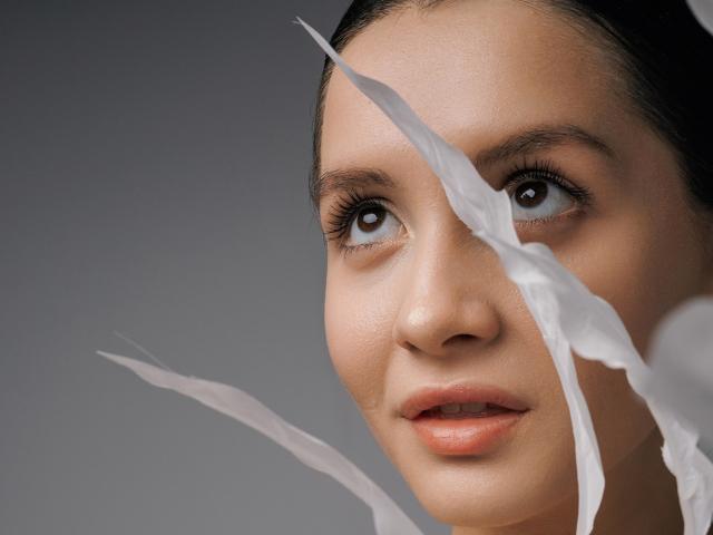 JeanieCat's face with white paper strips partially covering her eyes, showing her brown eyes and slight smile. She has fair skin and dark hair, looking up.