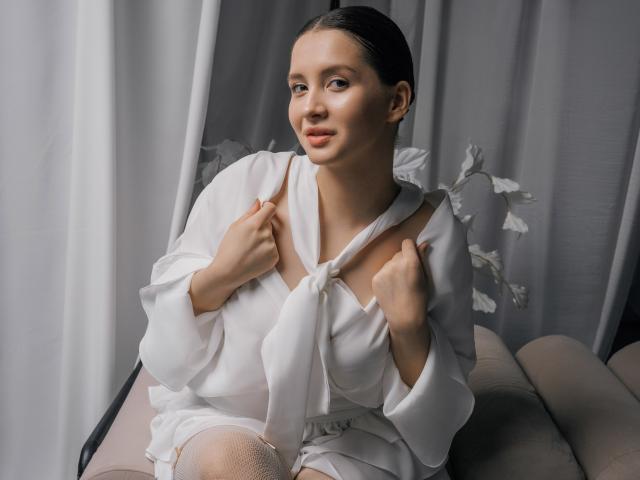 JeanieCat with fair skin, black hair, and soft makeup, wearing a white, loose blouse with a tied front, holding the fabric, sitting on a beige chair, smiling.