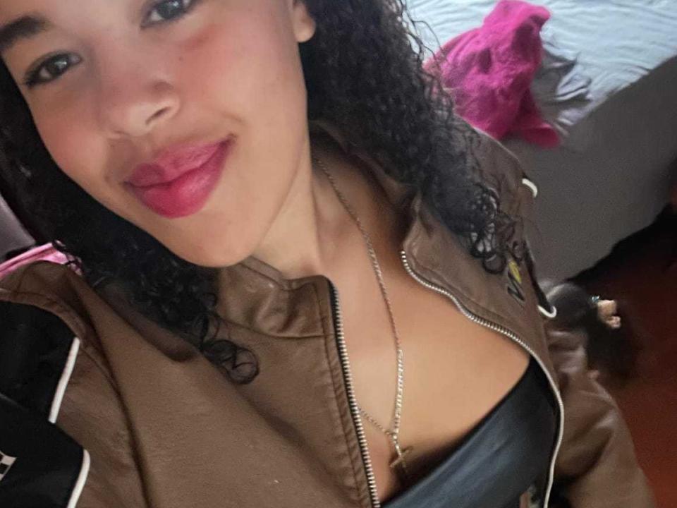 ZuriMontrel mit lockigem, schwarzem Haar, einer braunen Jacke und rotem Lippenstift, zeigt ihre Brüste in einem schwarzen Leder-Top und lächelt in die Kamera. Ihre Halskette hängt zwischen ihren Brüsten..