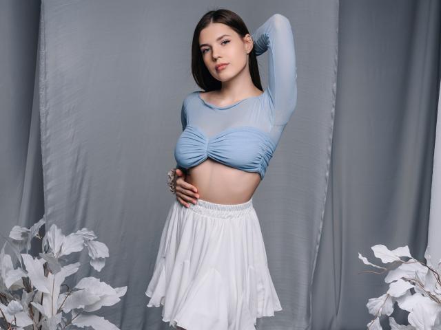BelleLovely with her tits up, wearing a sheer blue top and white skirt, hand on her belly, brown hair, and a subtle smile, against a gray backdrop.