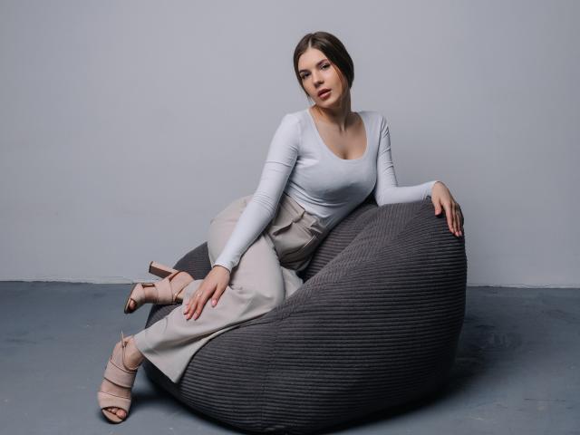 BelleLovely with tits in a tight white top, sitting on a gray ribbed beanbag, wearing beige pants and pink strappy heels, looking at the camera.
