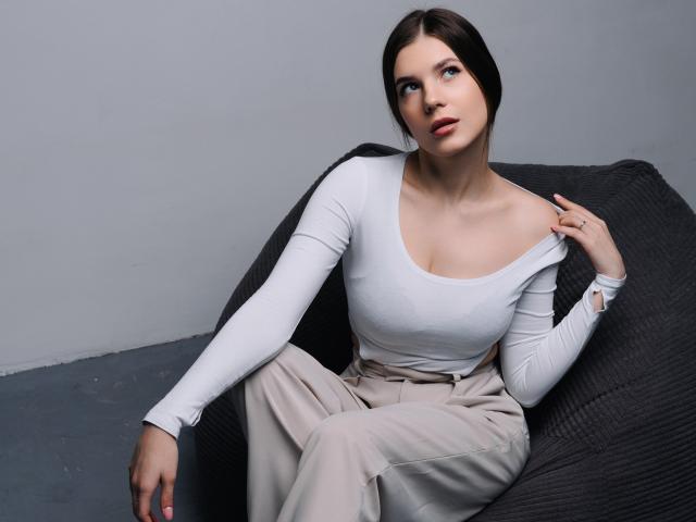BelleLovely with white long-sleeve top, showing her small tits, sitting on dark gray chair, beige pants, brown hair, light skin, hand on shoulder, looking up.