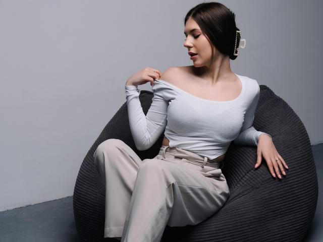 BelleLovely with small tits, off-shoulder white top, and beige pants, sits on a dark gray, textured chair, adjusting her top, with a subtle, confident smile.