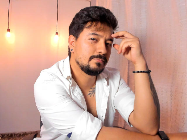 DavidEvans with short black hair and beard, white shirt, black bracelet, tattoo on chest, dark ear plugs, thinking with finger on cheek, light bulbs in background.