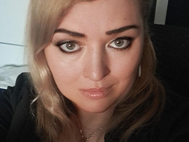 CamilleJazz has puffy, shiny lips, light brown eyes with thick eyeliner, blonde wavy hair, and wears small white earrings. Her nose is slightly upturned. She's wearing a black top.