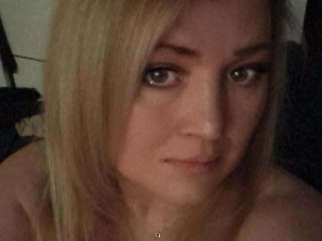 CamilleJazz has blonde hair, green eyes, and a slightly serious expression. Her bare shoulders are visible, and she's wearing a gold necklace. The photo is blurry.