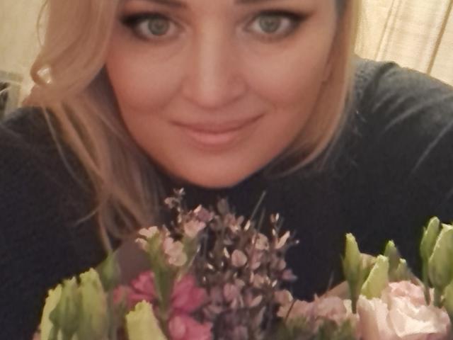 CamilleJazz with blonde hair, blue eyes, and black eyeliner, smiling at the camera, holding a bouquet of pink, white, and green flowers. Her tits are partially visible.