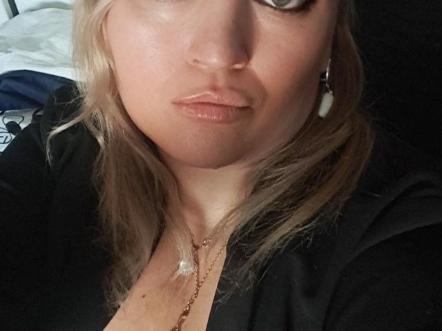 CamilleJazz's face with light blonde hair, wearing a black top, subtle makeup, and white earrings, showing a small, visible cleavage with a necklace. Her lips are slightly pursed.
