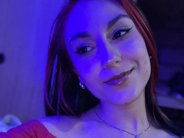 AmyStreetwise69 with red hair, blue neon light, eyeliner, piercing, glossy lips, and necklace, smiling slightly, showing a small amount of white fluid on her chin.