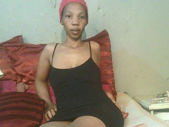Sexytinny-hot with brown skin, small tits, wearing a red beanie and black spaghetti-strap dress, sitting on red pillows, looking at the camera with a neutral expression.