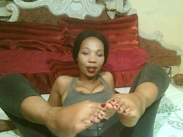 Sexytinny-hot with dark skin, short black hair, wearing a gray tank top and black pants, lying on a bed with legs up, showing bare feet and red toenails.