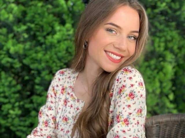 OliviaaDiamond mit langen braunen Haaren, einem Blumenkleid und rotem Lippenstift, lächelt in die Kamera. Sie hat blaue Augen und kleine Ohrringe. Ihre Brüste sind nicht sichtbar..