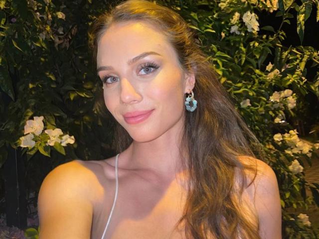 OliviaaDiamond tem olhos azuis, cabelos castanhos longos e está usando brincos verde-água com flores brancas. Ela está sorrindo com batom rosa, exibindo seus seios em uma blusa branca com alças..