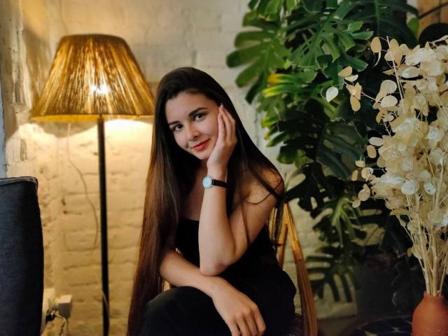 SofaNeumann, com cabelos castanhos longos, vestido preto, sorriso adorável, com o queixo apoiado na mão, uma luminária dourada, uma planta verde, flores brancas e um relógio de pulso, sentada em uma sala aconchegante..