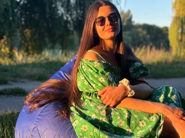 SofaNeumann, com cabelos castanhos longos, vestindo um vestido verde com estampas florais, óculos de sol e uma tiara branca, sentada em um pufe azul sob a luz do pôr do sol, com uma expressão relaxada e confiante..
