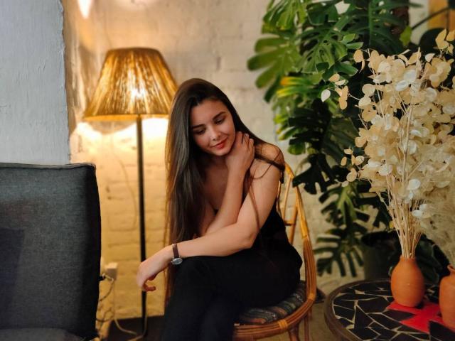 SofaNeumann avec de longs cheveux bruns, de petites seins, une robe noire, assise sur une chaise en osier, souriante, un bras sur la poitrine, une lampe, une plante verte, des fleurs blanches en arrière-plan, un canapé gris foncé..