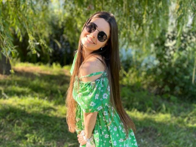 SofaNeumann com cabelos castanhos longos, vestido floral verde, com um ombro descoberto, sorrindo, usando óculos de sol escuros, em pé na grama iluminada pelo sol, com as mãos nos quadris, olhando por cima do ombro, feliz, fofa..