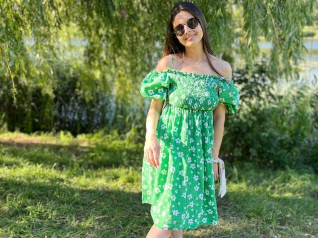 SofaNeumann is wearing a green dress with white floral patterns, off-shoulder sleeves, and dark sunglasses. She's holding a white purse, standing in a grassy area.