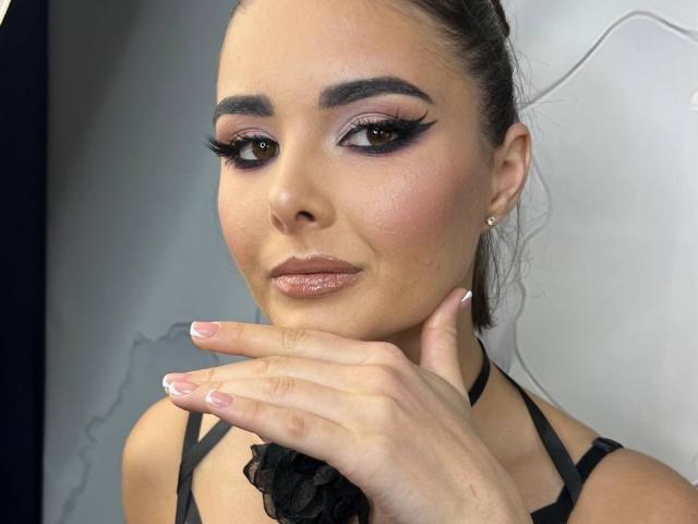 SofaNeumann met donkere wenkbrauwen, lange wimpers, roze oogschaduw, glanzende bruine lippen, een zwarte choker en roze nagellak, hand in de buurt van het gezicht, kijkend naar de camera..