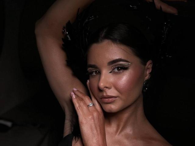 SofaNeumann, avec ses cheveux foncés, un chapeau orné de plumes noires, portant une bague en diamant, posant les bras levés, avec une peau lumineuse, un maquillage subtil, et une expression confiante et séduisante..