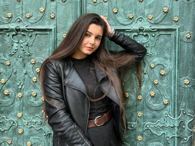 SofaNeumann met lang bruin haar, een zwarte leren jas en een bruine riem, staat tegen een groene, rijkelijk versierde muur, met haar hand in haar haar, zelfverzekerd, sexy en stijlvol..