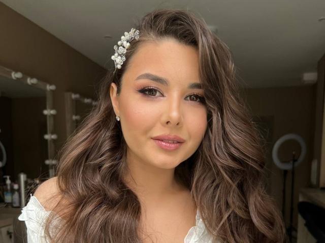 SofaNeumann has wavy brown hair, a flower hairpiece, pink eyeshadow, subtle makeup, pink lipstick, and small diamond earrings. She's smiling with a cute, sexy look.